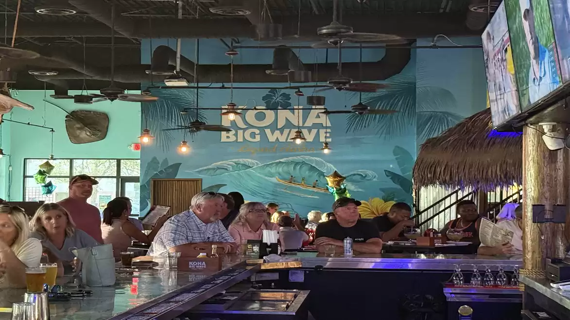 A lively interior of a beach bar with patrons sitting at the counter and a mural with the text 'Kona Big Wave'.
