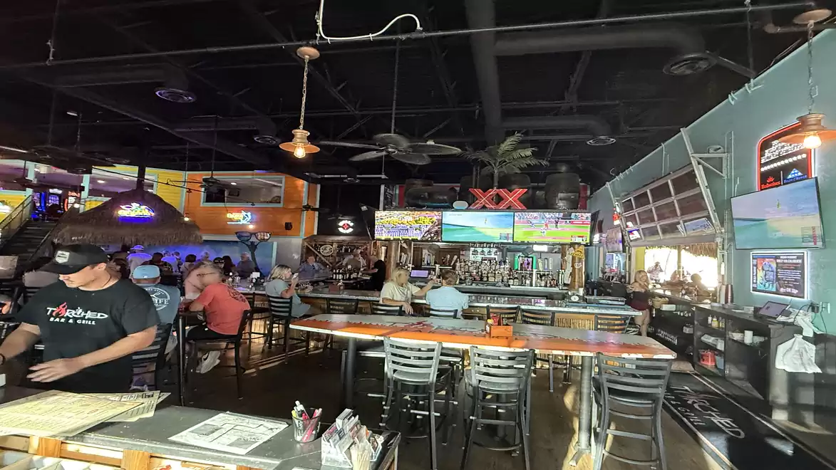 Interior view of a busy bar with colorful lit ceilings and screens displaying sports broadcasts. People are seated at tables and the bar is centrally located.