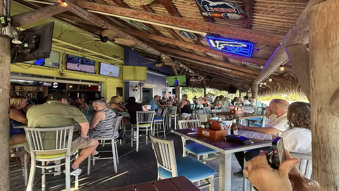A busy open-air restaurant with a thatched roof, where people sit on bar stools and at tables enjoying a relaxed atmosphere.