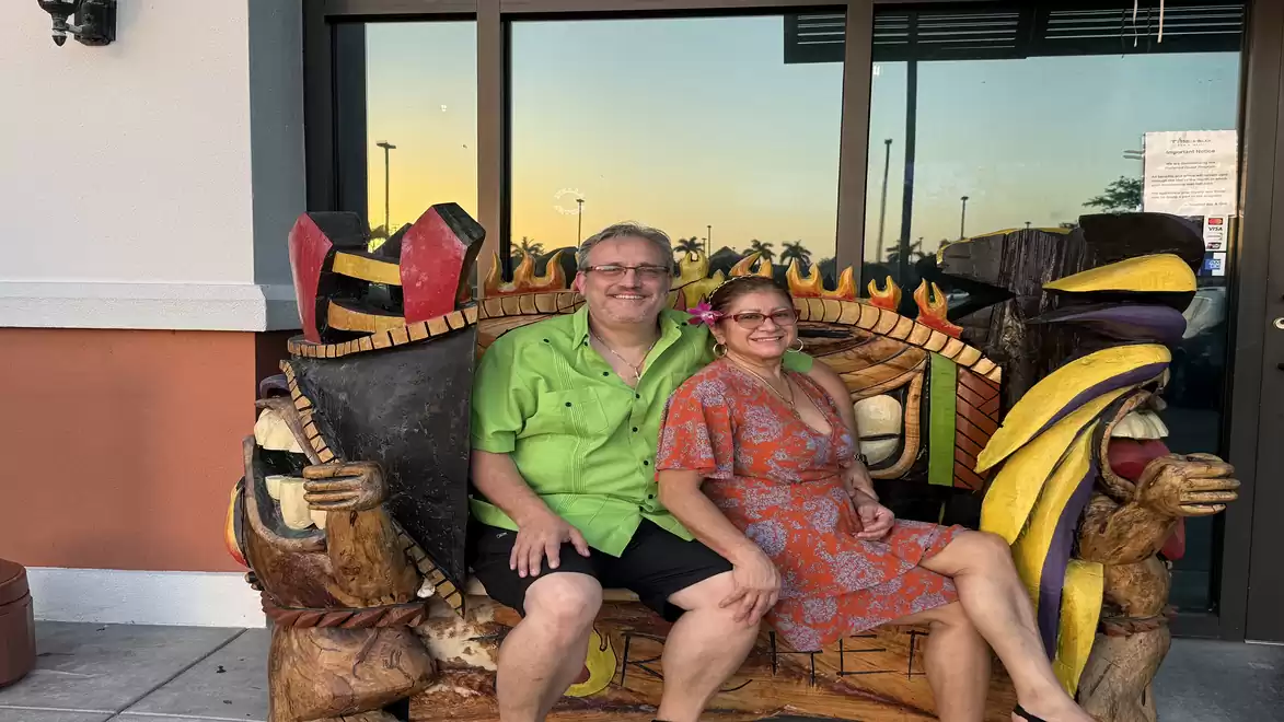 A smiling couple sits on a decorative, colorful wooden bench in front of a building at sunset.
