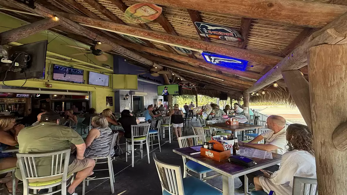 A cozy beer garden with a thatched roof with many people sitting at tables watching football on several TVs.