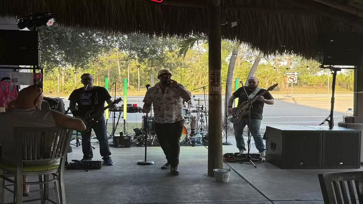 A band performing live on a covered event space. The singer is in the front, with two guitarists and a drummer playing in the background. The scene is illuminated by daylight.