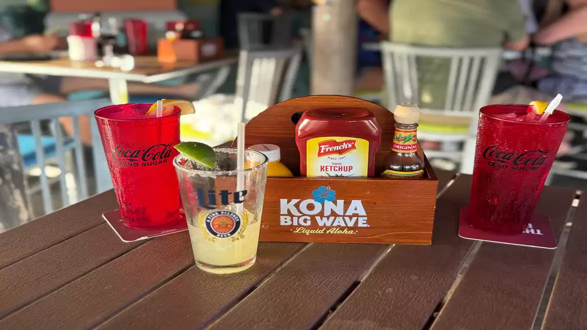 A table in a restaurant with drinks, condiments, and a box. Two red drinks in Coca-Cola glasses flank a yellow drink with lime. In the center is a condiment box with ketchup and other sauces.