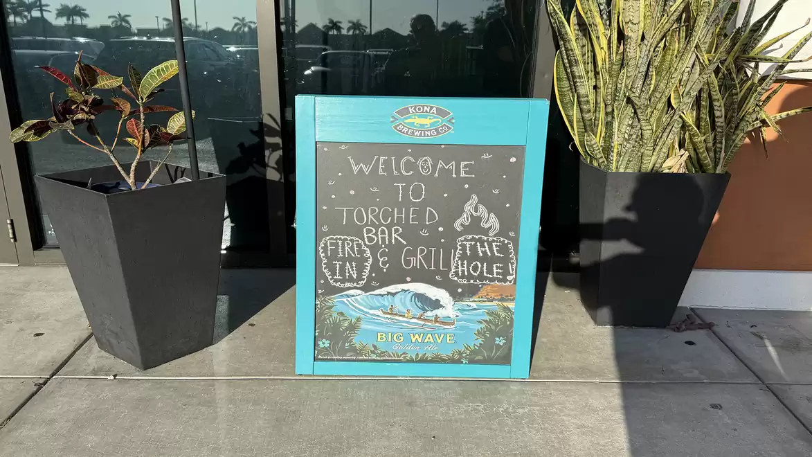 A blue-framed sign in front of a restaurant with the text 'Welcome to Torched Bar & Grill' next to two large plant pots.