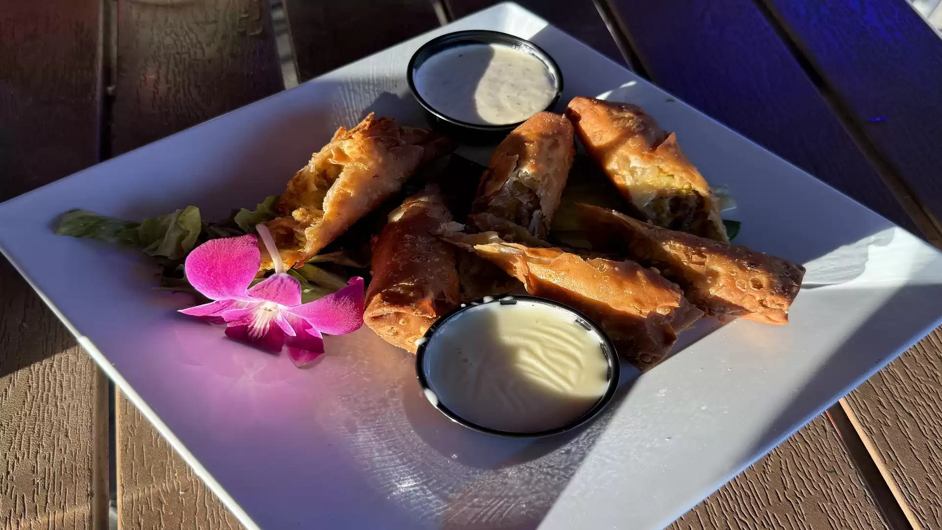 Close-up of a plate with spring rolls and two dips, garnished with a pink flower on a white plate.