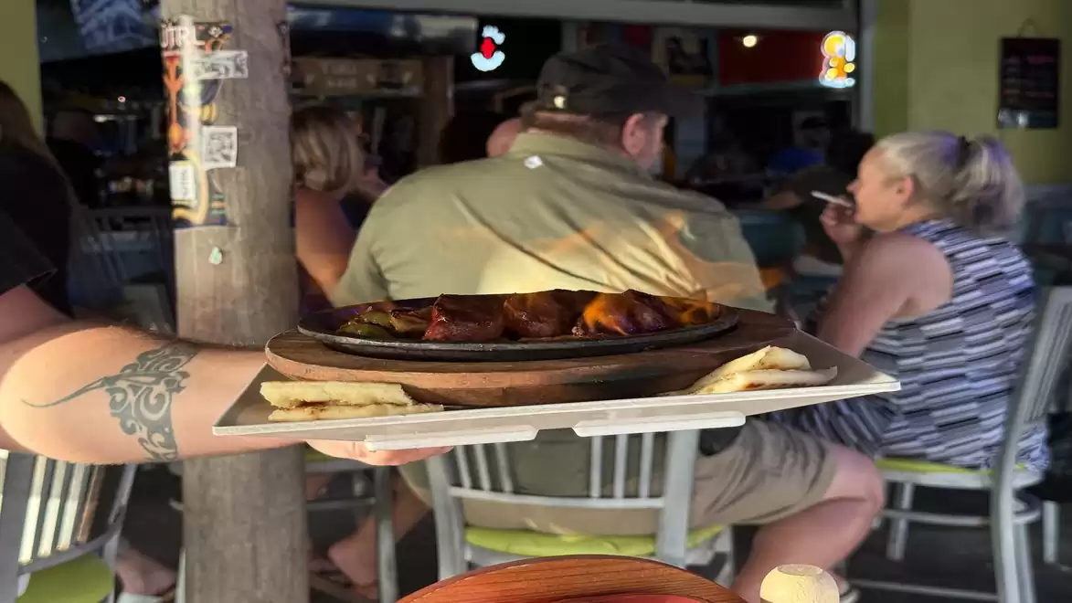 A waiter serves a hot dish on a metal plate in a busy restaurant. In the background, guests are seen sitting at tables.