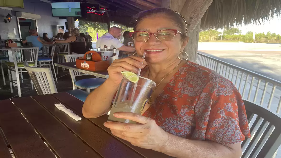 A woman sits in an outdoor restaurant drinking from a glass with a straw. She is wearing glasses and smiling at the camera.