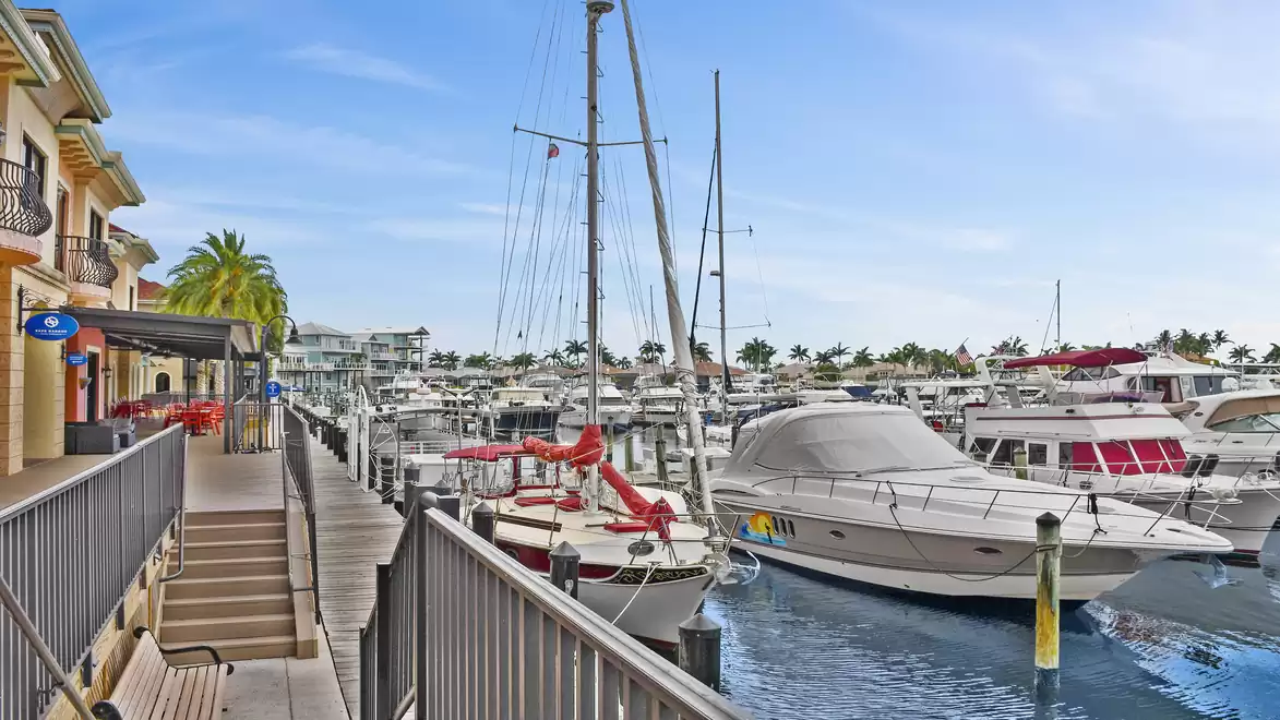 A modern marina with a variety of luxurious boats and yachts in clear blue water. In the foreground, there is a walkway with railings and palm trees surrounding the harbor.