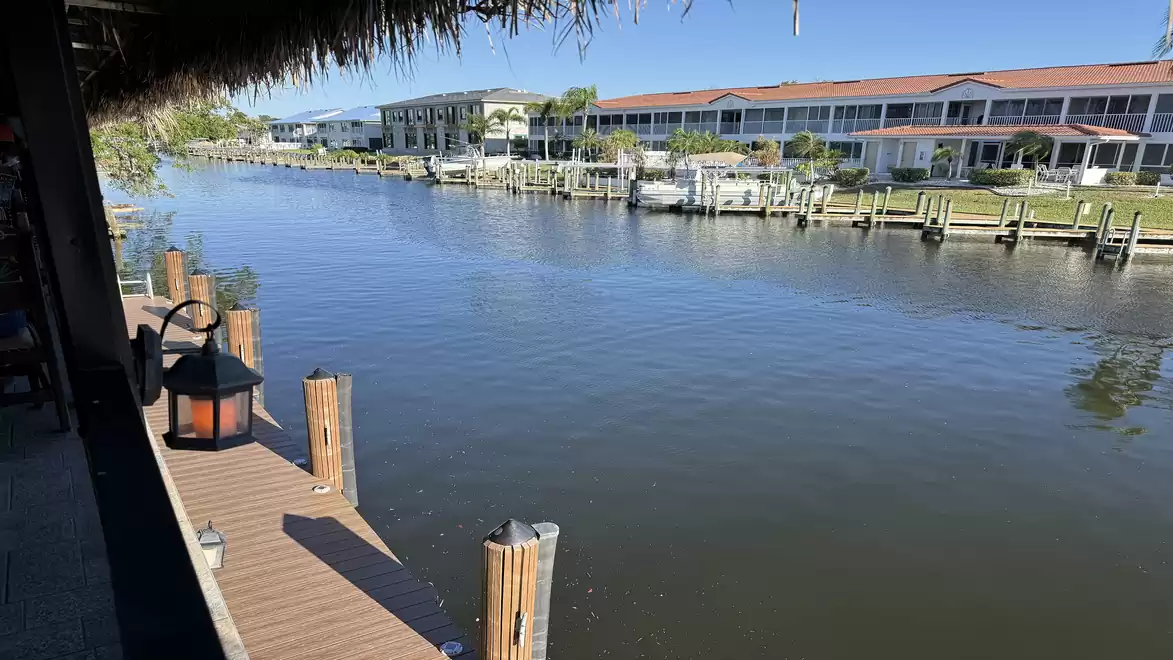 Image of a calm canal with a dock, lamps, and residential buildings in the background on a sunny day.