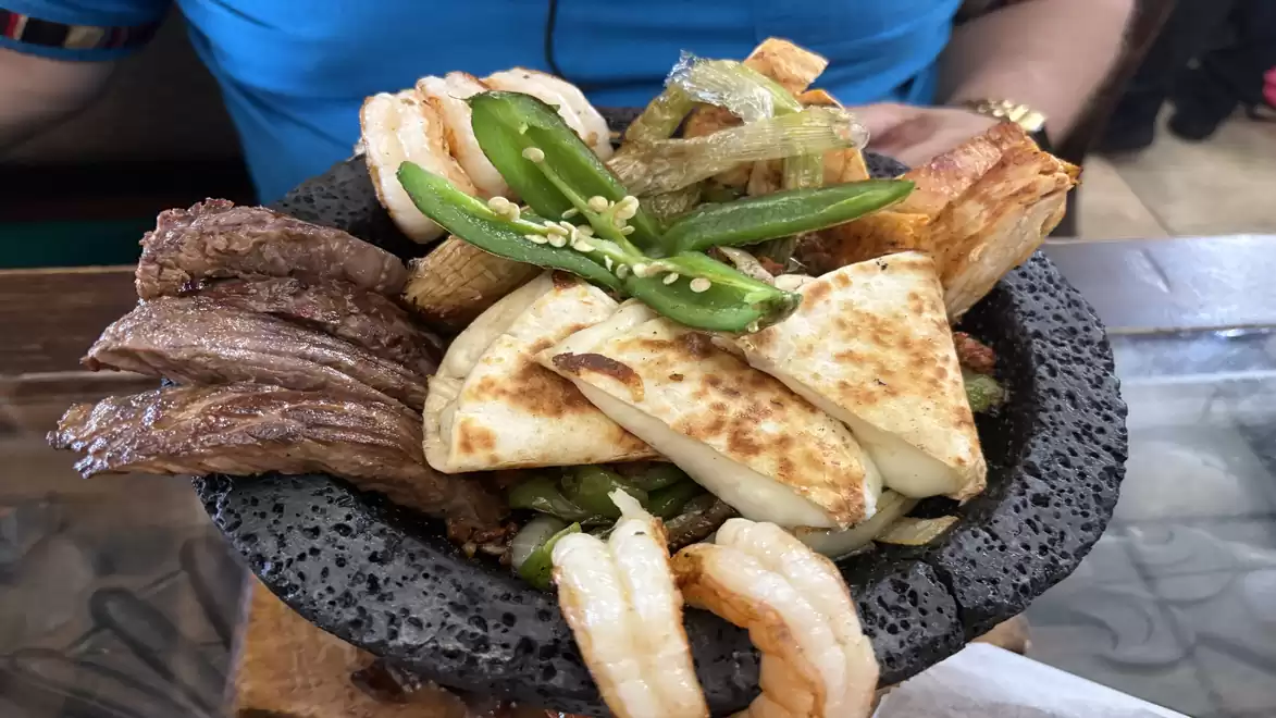 A hearty dish featuring grilled meat, shrimp, vegetables, and grilled cheese, served on a black rustic plate.