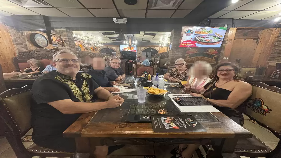 A group of people sitting together at a wooden table in a restaurant. Decorative elements and a TV are visible in the background.
