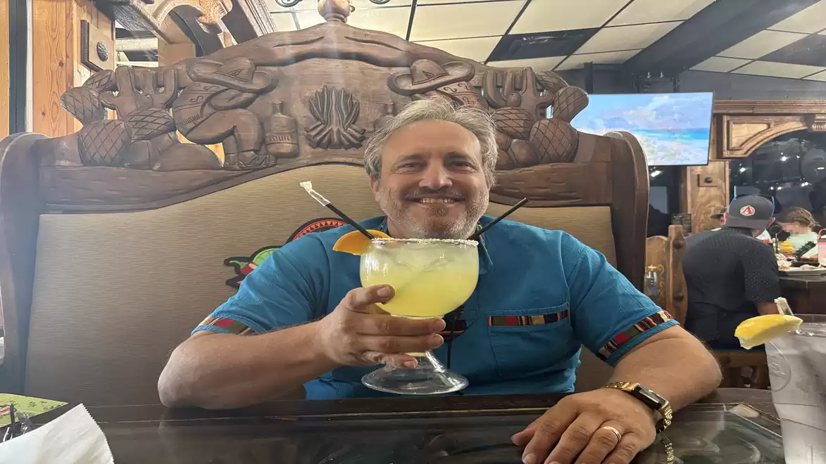 Man in a blue shirt holding a large glass with a yellow drink in a restaurant with wooden carvings in the background.