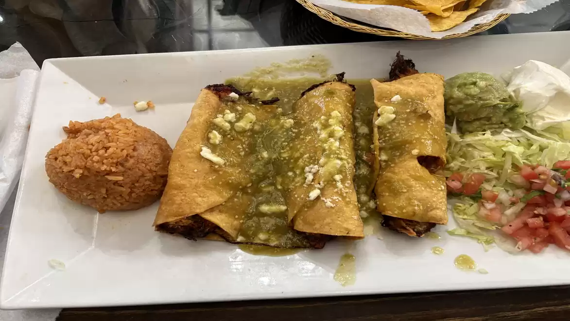 A plate with Mexican dishes, including three filled tortillas with green salsa, a dollop of guacamole, rice, and a salad of tomatoes and lettuce.