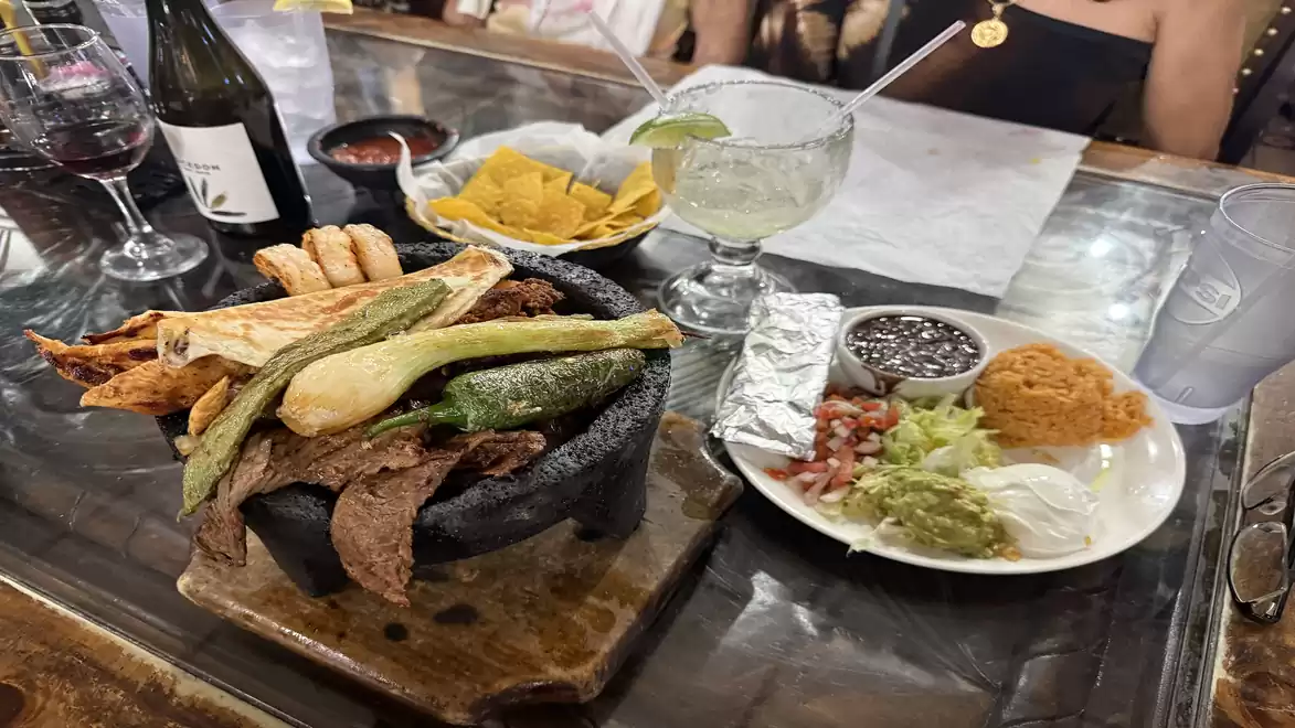 A table with Mexican dishes including grilled meat and vegetables on a stone platter, accompanied by a plate with sides like beans, guacamole, and rice, next to chips and drinks.