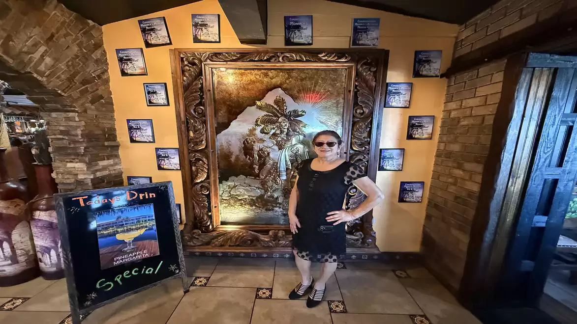 A person stands in a room with a large, ornately framed wall decoration, surrounded by smaller wall pictures. On the left, there is an advertisement board for a drink special.