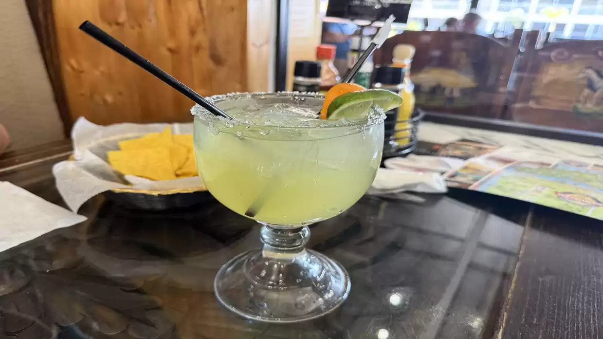 A refreshing drink in a glass with a black straw, garnished with lime and orange, on a table in a restaurant. Tortilla chips and various sauce bottles are visible in the background.