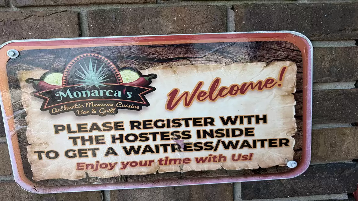 A sign on a brick wall advertising Monarca's Bar & Grill restaurant. The text instructs to register with the hostess inside to get service from a waiter or waitress.