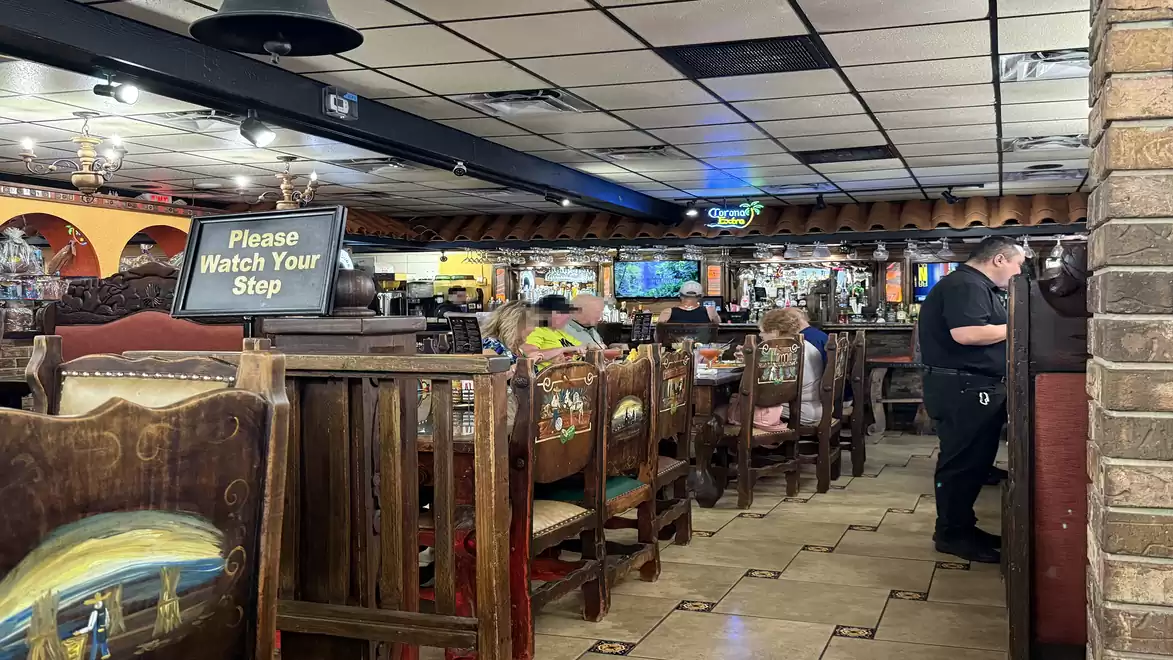 Interior view of a rustic restaurant with wooden furniture and decorative elements. Guests are seated at tables enjoying their meals. A sign saying 'Please Watch Your Step' is visible.