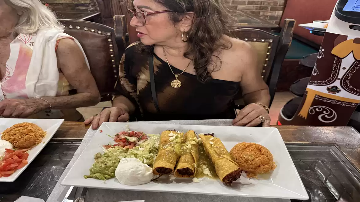 A woman sits at a table with Mexican food in front of her, including taquitos, rice, guacamole, and sour cream.
