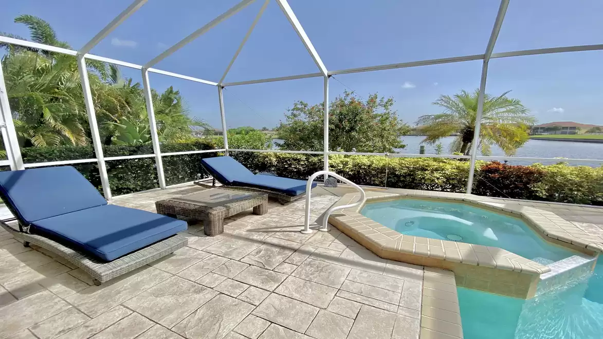 Pool area with two blue loungers, a table, and a hot tub. Plants and a lake are visible in the background.