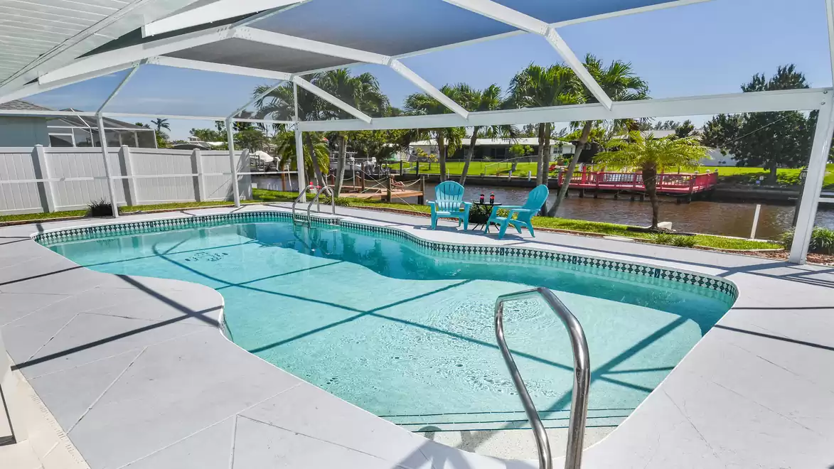Villa Coral Sunshine - Modern outdoor pool with canopy, surrounded by white fence and palm trees. In the background, a water canal and two blue chairs with a table.