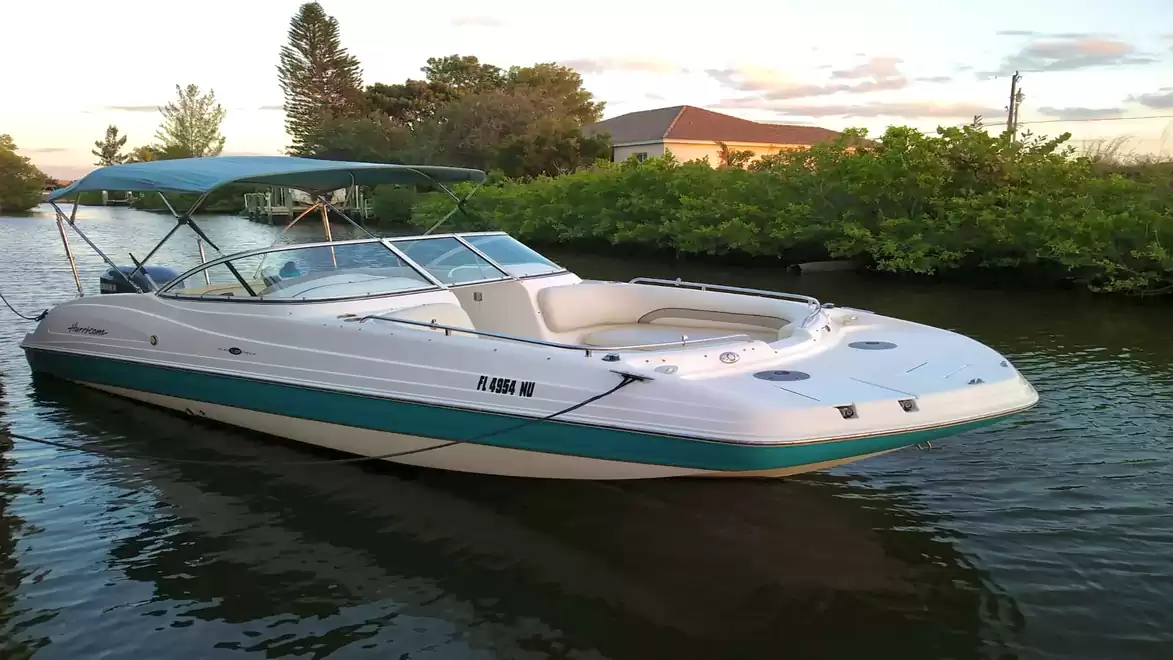 A white motorboat with a green stripe is cruising on a river or canal, surrounded by trees and a house in the background under a slightly cloudy sky.
