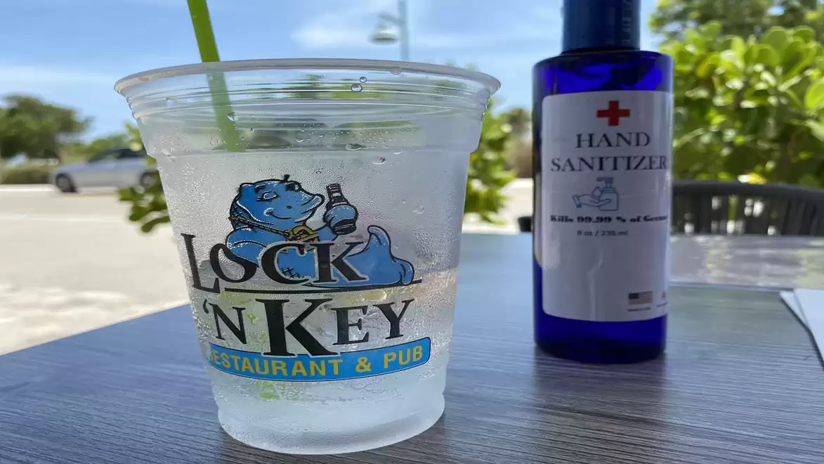 A plastic cup with a logo of a blue hippopotamus and the text 'Lock and Key Restaurant & Pub' sits on an outdoor table. Next to it is a blue bottle of hand sanitizer.