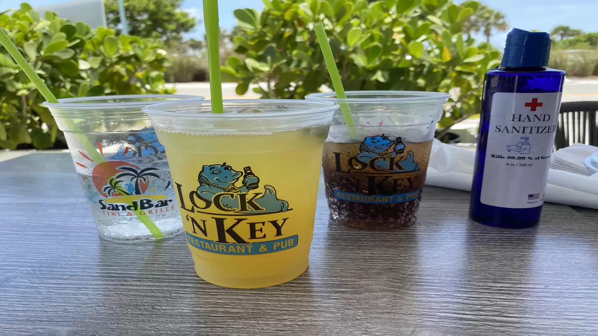 Four plastic cups with drinks and a hand sanitizer on a table in an outdoor setting with plants in the background.