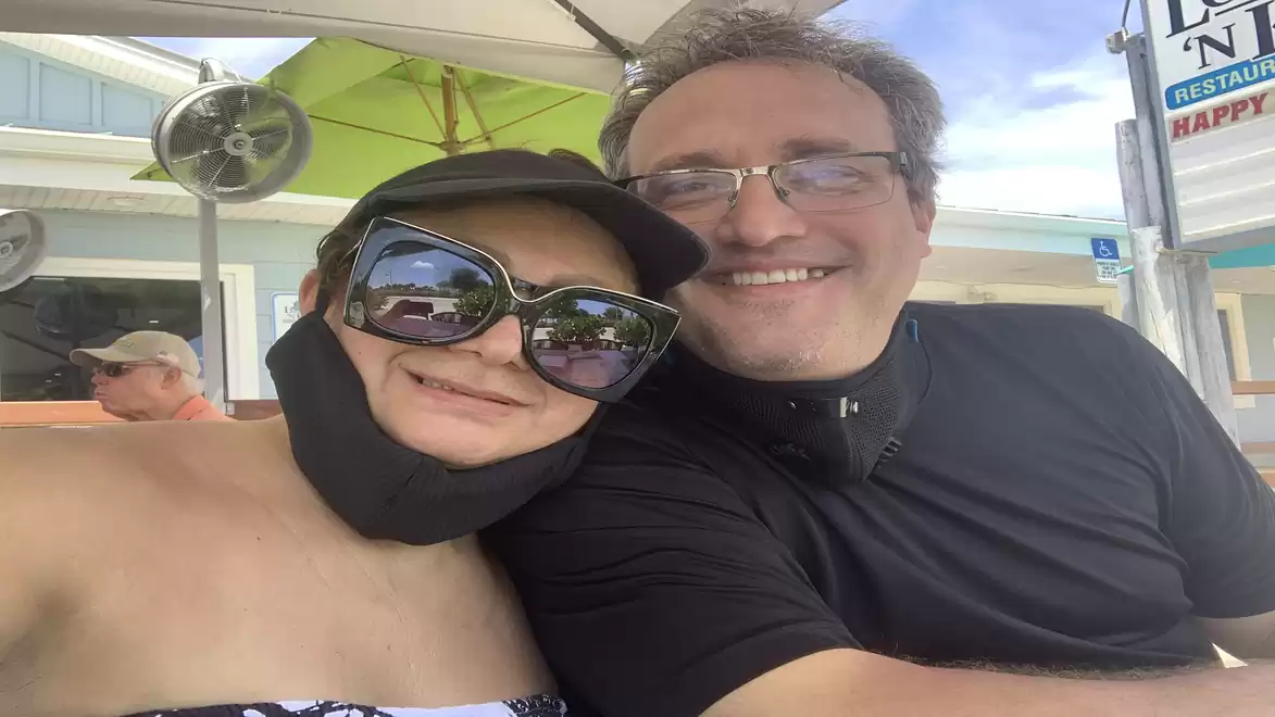A man and a woman in summer clothing and large sunglasses smile at the camera. They are sitting outdoors under a parasol at a table.