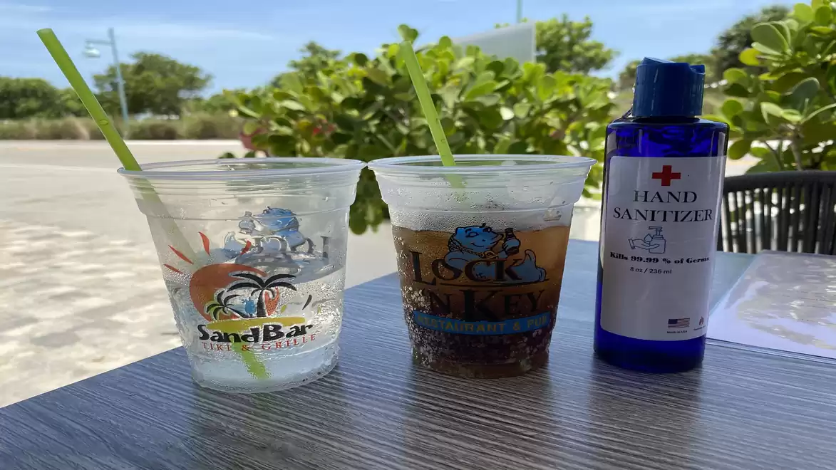 An outdoor table with two plastic cups, one with a clear drink and one with a dark drink. Next to the cups is a bottle of hand sanitizer. Green bushes and a sunny sky are visible in the background.