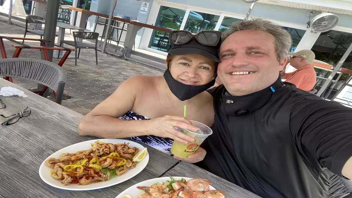 A smiling couple is sitting in a restaurant. In front of them are two plates with food and a drink. They are wearing casual clothing, and sunglasses are visible.