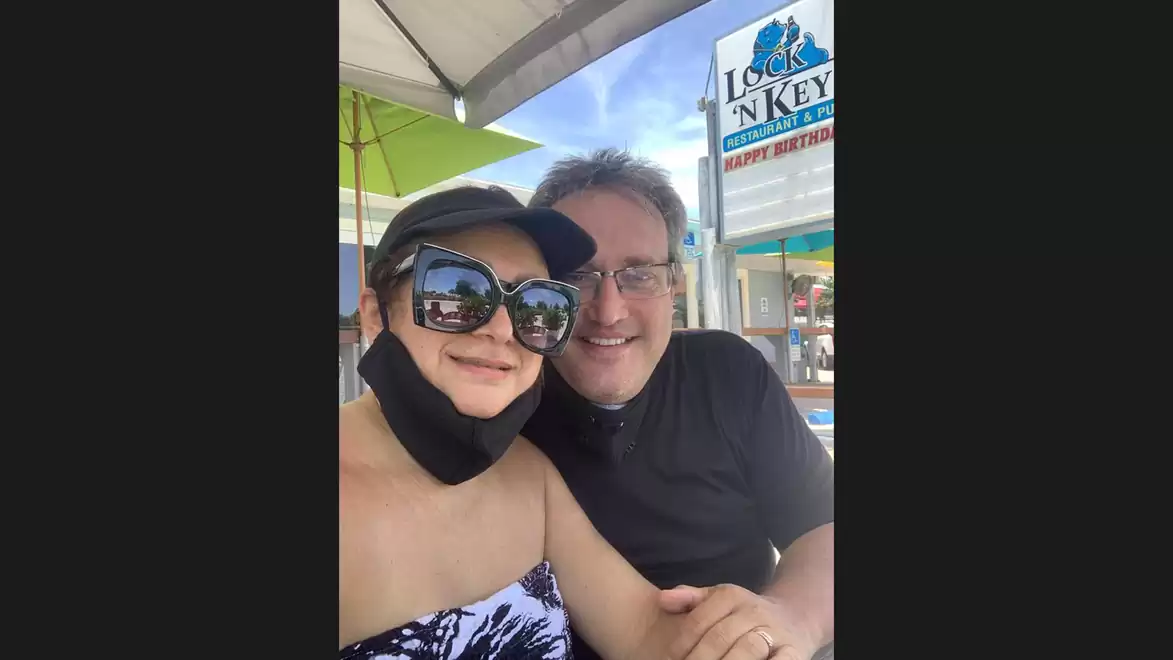 A smiling couple sitting outdoors under a sun umbrella. The woman is wearing sunglasses and a black dress, the man a black T-shirt. A sign is visible in the background.