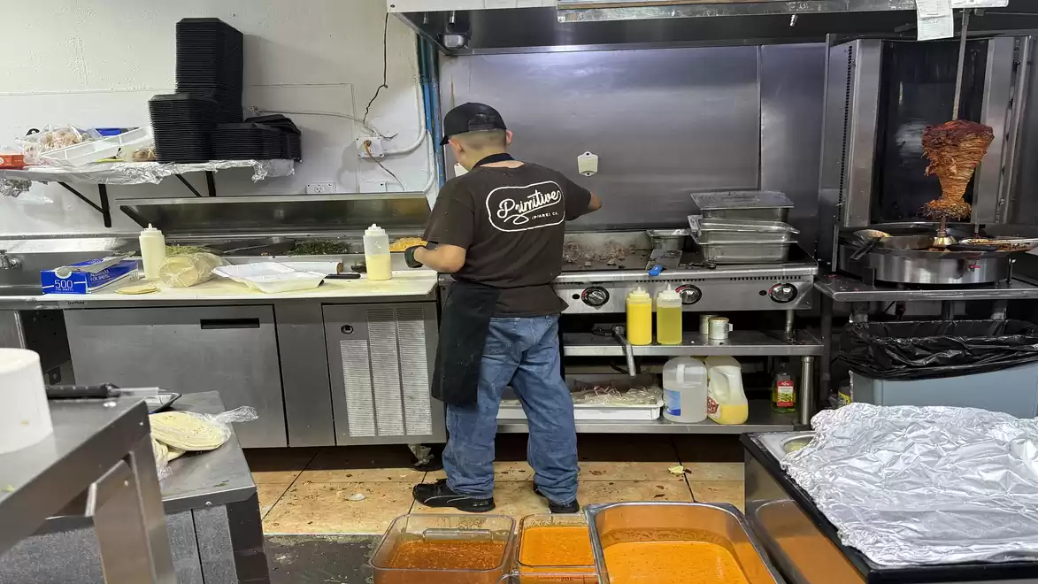 A chef working in an industrial kitchen with stovetops, a kebab grill, and various ingredients.