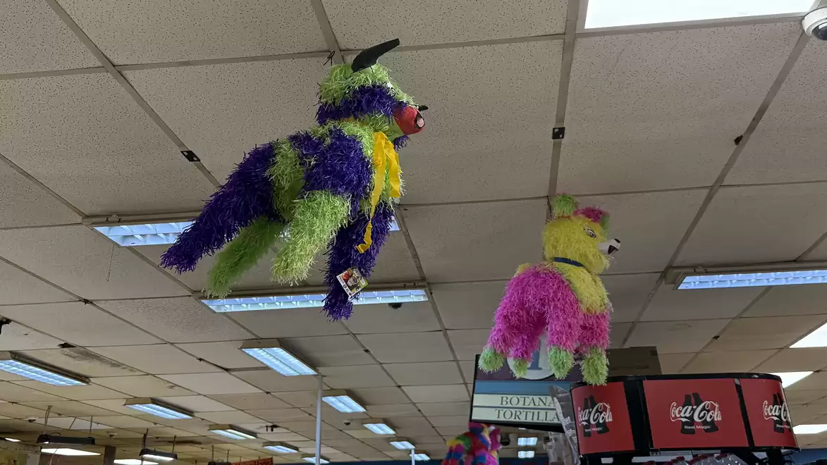 Two colorful animal-shaped pinatas hanging from the ceiling of a supermarket. One pinata is purple and green, the other is pink and yellow.