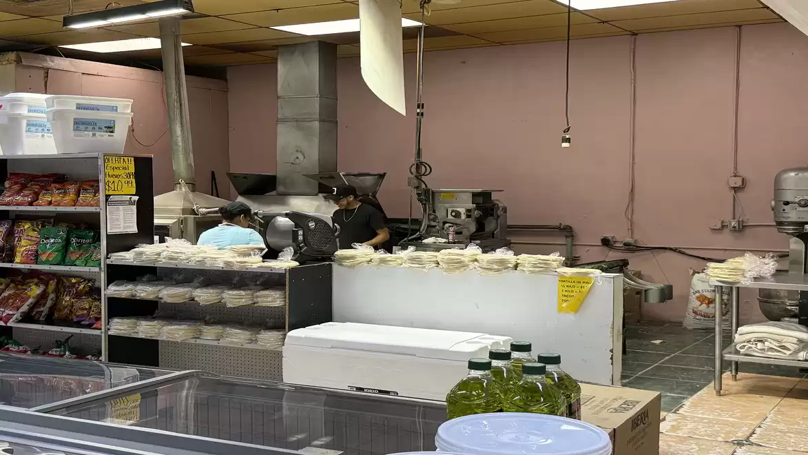 A deli in a kitchen with two people working at a counter with food. In the background, shelves with snacks and industrial kitchen equipment are visible.