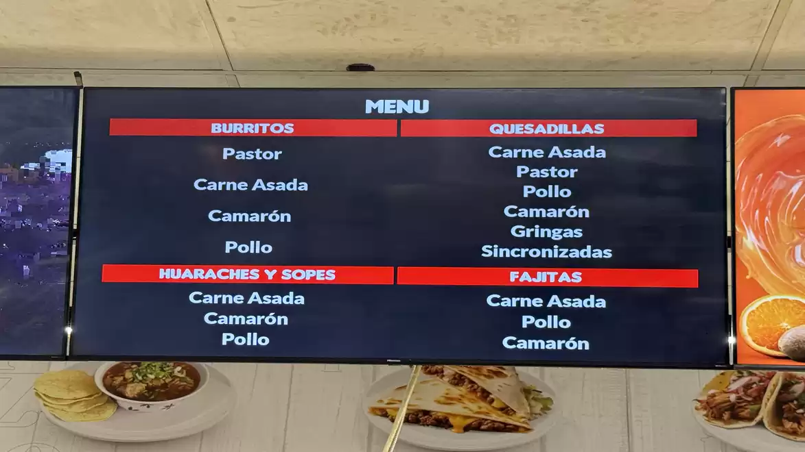A digital menu board in a restaurant displays various Mexican dishes like Burritos, Quesadillas, Huaraches, Sopes, and Fajitas with meat options like Pastor, Carne Asada, Camarón, and Pollo.
