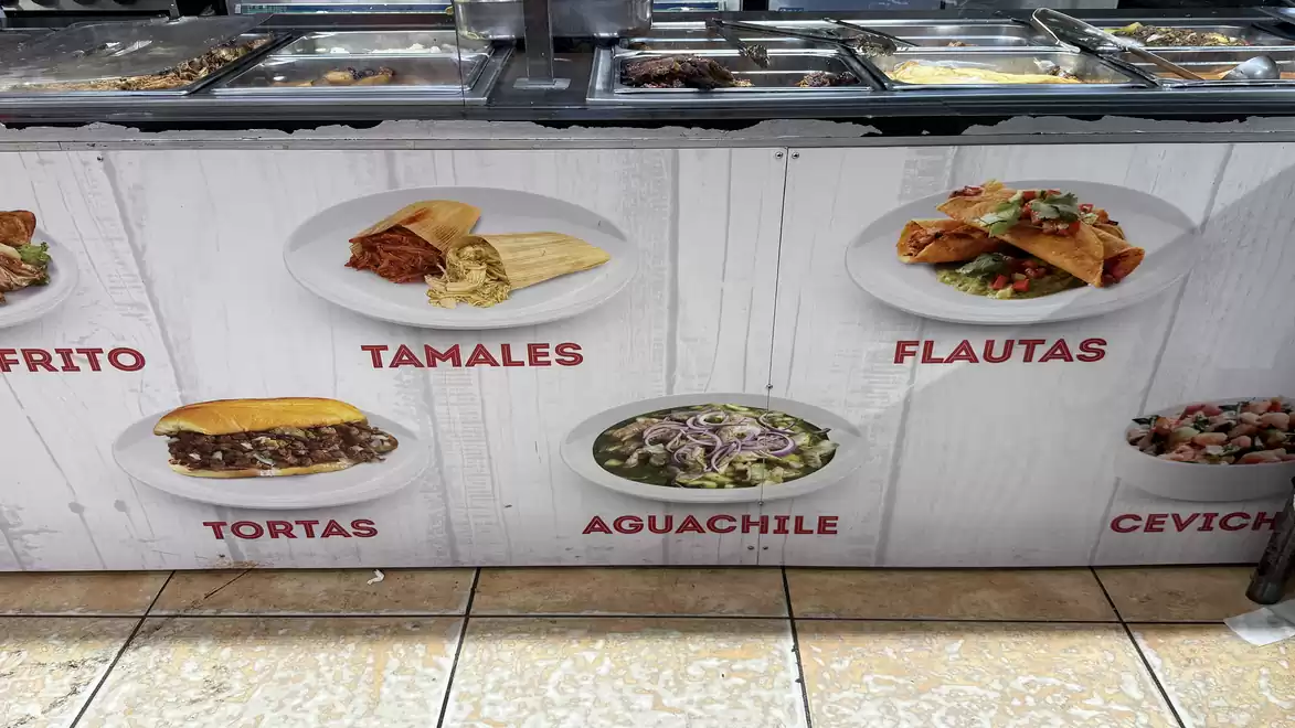 Photo of a food stand displaying various dishes: Elote Frito, Tamales, Flautas, Tortas, Aguachile, and Ceviche.