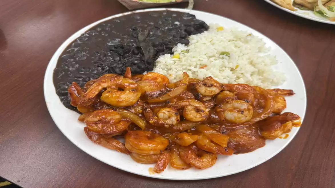 A plate of fried shrimp in sauce, served with white rice and black beans on a wooden table.