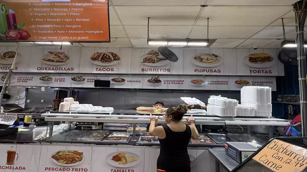 A woman stands at a food stall in a market hall ordering from an employee. Behind the counter, signs display various Mexican dishes like quesadillas, tacos, burritos, and more. A stack of styrofoam containers is prominently visible.