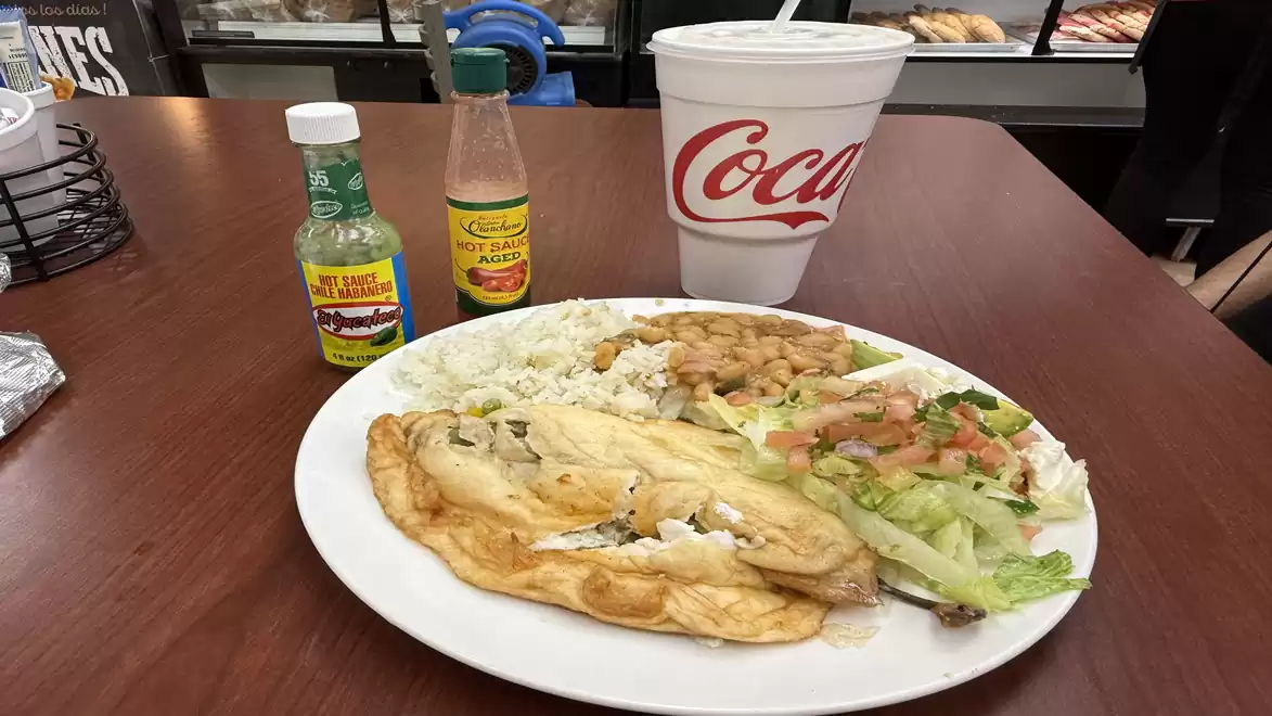 A plate of Mexican food, including a filled tortilla, rice, and salad, on a wooden table with two bottles of sauce and a large Coca-Cola cup.