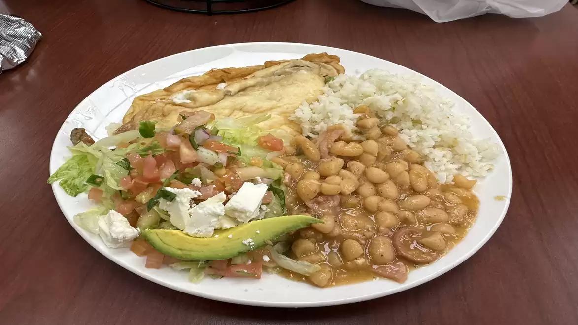 A plate with rice, beans, salad with tomatoes and feta, a slice of avocado, and a piece of baked chicken.