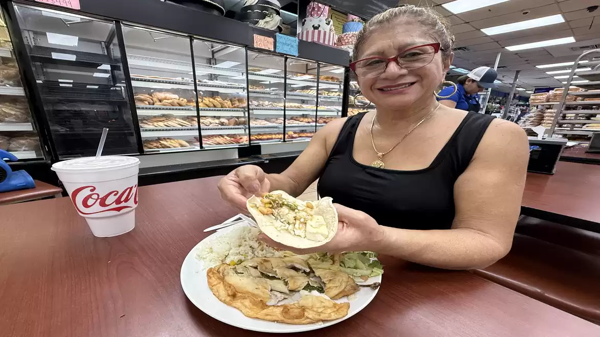 A smiling woman sits at a table in a bakery or snack bar, holding a filled taco. Various foods are on her plate, and next to it is a drink cup with Coca-Cola.