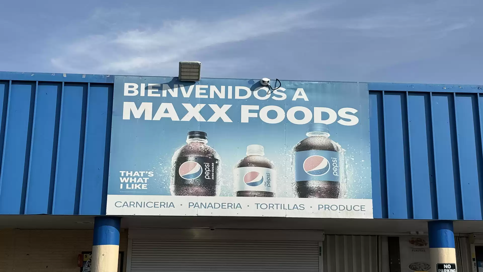 Advertising sign of a supermarket with beverage bottles and the text 'Bienvenidos a Maxx Foods'. The blue background and Pepsi logos are prominent.