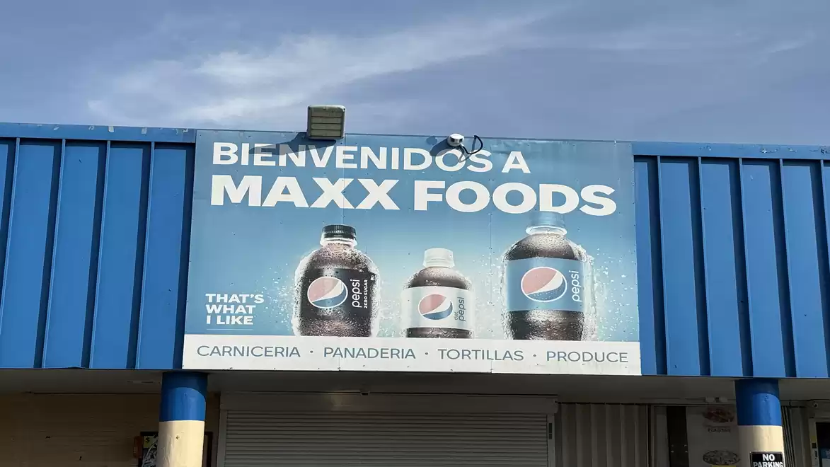 Advertising sign of a supermarket with beverage bottles and the text 'Bienvenidos a Maxx Foods'. The blue background and Pepsi logos are prominent.