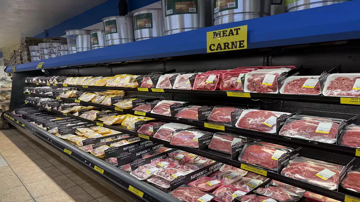 Refrigerated shelf with various packaged meats in a supermarket, including poultry and red meat, with a 'Meat Carne' sign above.