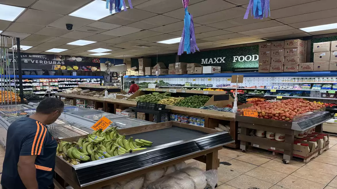 A well-stocked supermarket with fresh vegetables and fruits, colorful decorations on the ceiling, and a customer shopping.