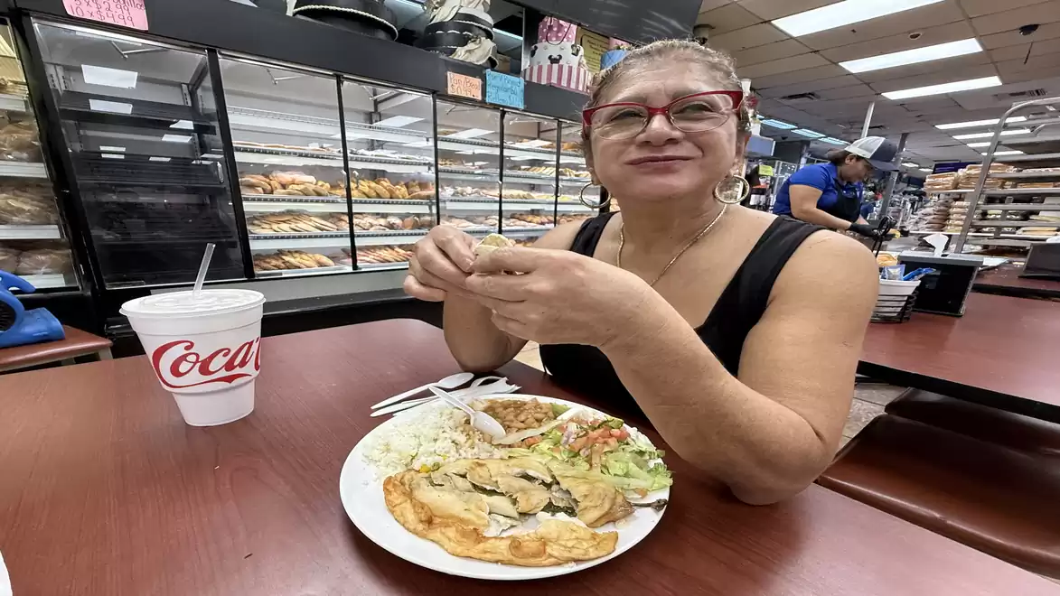 A woman sits in a café or bakery enjoying a meal with rice, vegetable mix, and omelette at a table with a drink cup. Various bakery items are visible in the background.