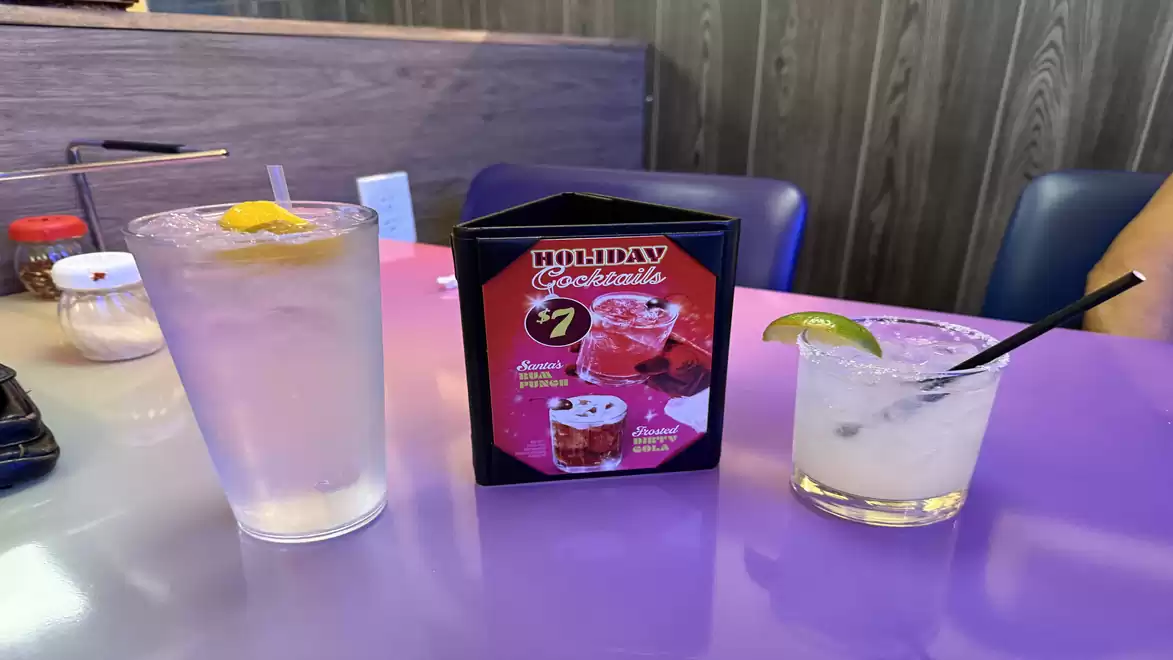Three drinks on a table in a restaurant: A glass of water with lemon, a cocktails menu, and a cocktail with a lime slice.