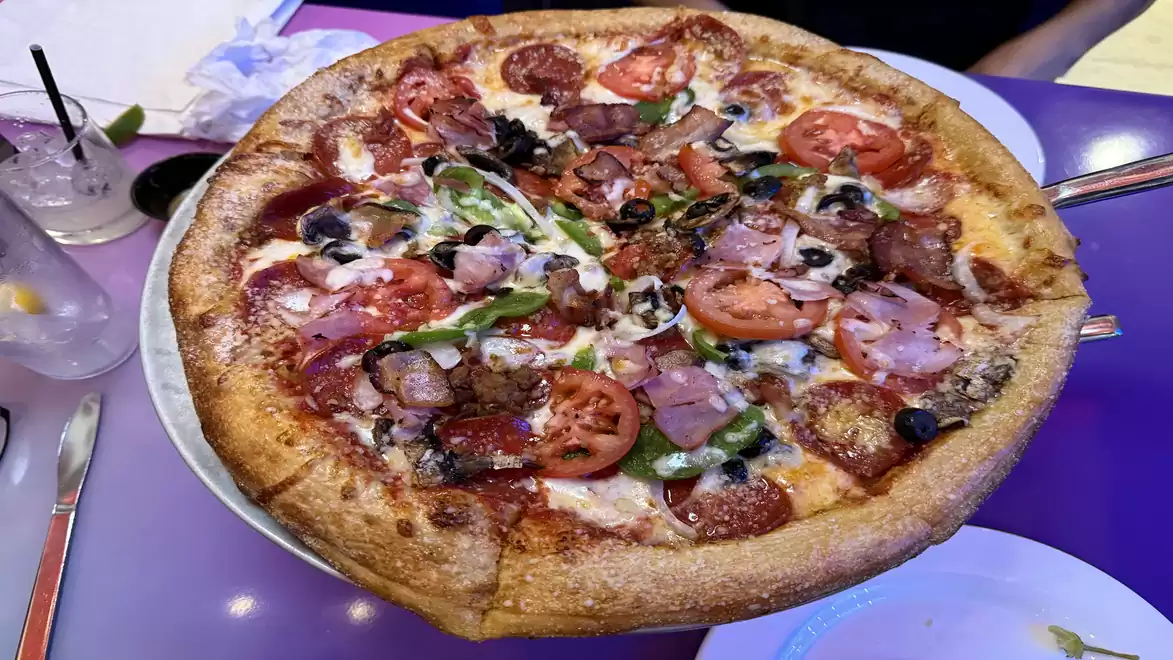 Close-up of a large freshly baked pizza with various toppings including tomatoes, olives, peppers, ham, and sausage on a purple table.