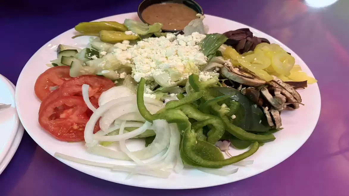 A plate with fresh salad consisting of tomatoes, onions, bell peppers, pickled peppers, cucumbers, mushrooms, olives, and feta cheese, with a dip on the side.